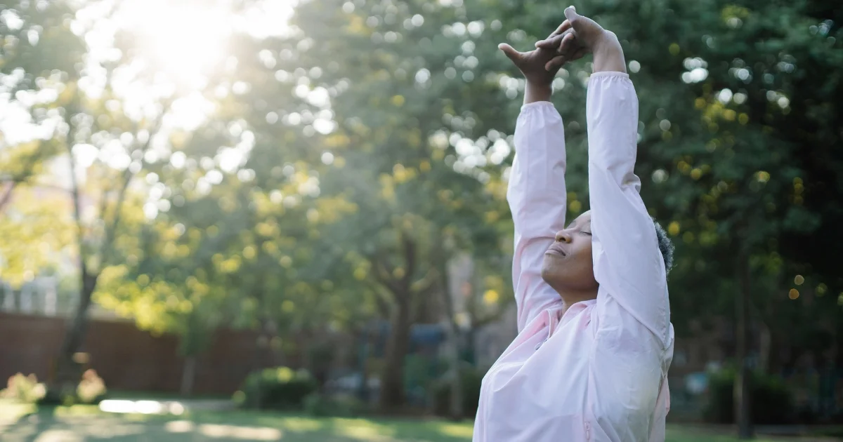 Woman in a pink jacket stretching with arms overhead in a sunny park setting.